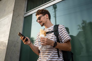 Genç, yetişkin, beyaz bir erkek, güneşli bir günde şehirde sandviç yiyor, fast food konsepti yiyor, şehir yaşamının fotokopisini çekiyor, turistleri duvarın önünde yiyor, gerçek bir insan cep telefonu kullanıyor.