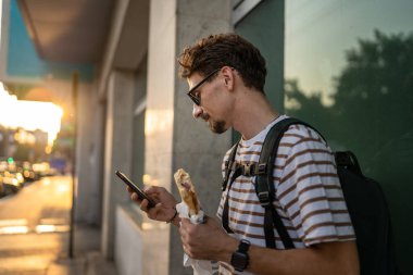 Genç, yetişkin, beyaz bir erkek, güneşli bir günde şehirde sandviç yiyor, fast food konsepti yiyor, şehir yaşamının fotokopisini çekiyor, turistleri duvarın önünde yiyor, gerçek bir insan cep telefonu kullanıyor.