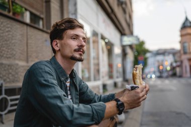 Genç, yetişkin, beyaz bir erkek, güneşli bir günde şehirde duruyor ve sandviç yiyor. Fast food konsepti. Şehir hayatı.