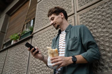 Gündüz vakti şehirde genç, yetişkin, beyaz bir erkek sandviç yiyor, fast food konsepti yiyor, sokak ortasında yer alan turistlerin fotokopisini çekiyor, gerçek bir insan cep telefonu mesajları kullanıyor.