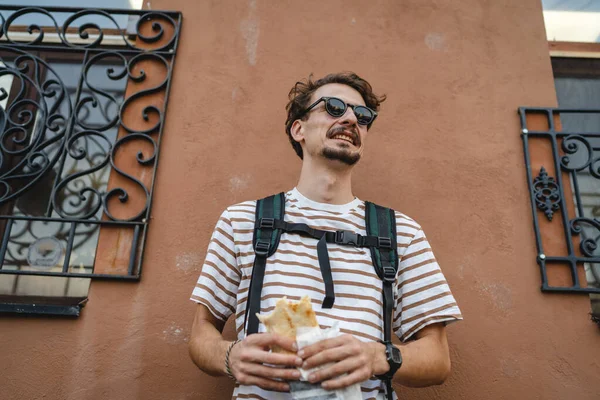 晴れた日のスタンドで市内の1人の若い大人の現代的な白人男性とサンドイッチを食べるファーストフードのコンセプト都市生活コピースペース観光客は壁の前で食べるリアル人の肖像画 — ストック写真