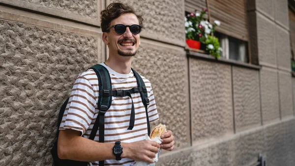 晴れた日のスタンドで市内の1人の若い大人の現代的な白人男性とサンドイッチを食べるファーストフードのコンセプト都市生活コピースペース観光客は壁の前で食べるリアル人の肖像画 — ストック写真