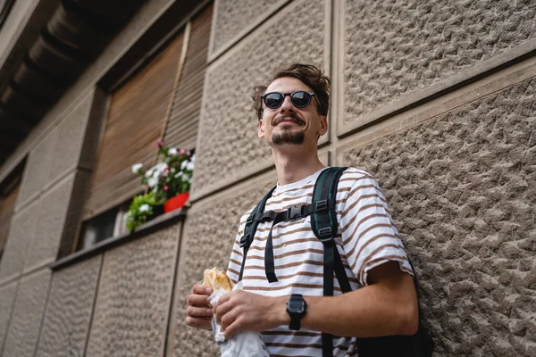 晴れた日のスタンドで市内の1人の若い大人の現代的な白人男性とサンドイッチを食べるファーストフードのコンセプト都市生活コピースペース観光客は壁の前で食べるリアル人の肖像画 — ストック写真