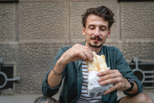 晴れた日のスタンドで市内の1人の若い大人の現代的な白人男性とサンドイッチを食べますファーストフードのコンセプト都市生活コピースペース観光客は壁の前で食べる本物の人 — ストック写真