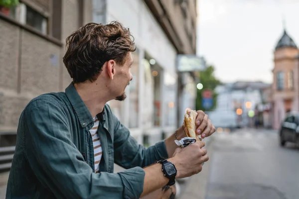晴れた日のスタンドで市内の1人の若い大人の現代的な白人男性とサンドイッチを食べますファーストフードのコンセプト都市生活コピースペース観光客は壁の前で食べる本物の人 — ストック写真