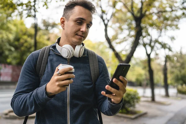 公園近くの街を歩いている若い大人の白人男性が携帯電話のスマートフォンを使ってSmsやビデオ通話をしています コーヒーを飲みながら幸せな男性観光客が立っています — ストック写真