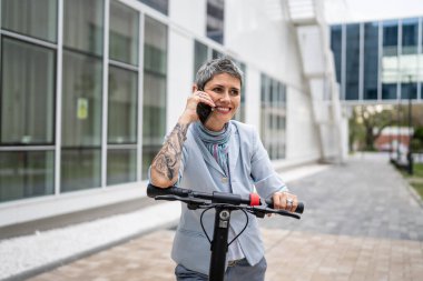 Yetişkin bir yaşlı beyaz kadın dışarıda duruyor. Cep telefonu kullanıyor. Gündüz güvenli modern alternatif ulaşım ve iletişim alanı kopyalama yönteminde elektrikli scooterla konuşuyor.