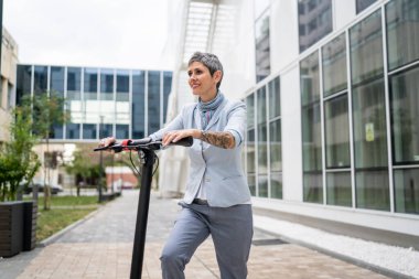 Bir kadın olgun, yaşlı, beyaz, açık havada elektrikli scooterlı bir kadın. Gündüz kendine güvenen bir iş kadını. Alternatif ulaşım yöntemini kullanıyor.