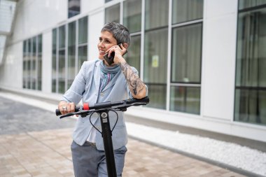 Yetişkin bir yaşlı beyaz kadın dışarıda duruyor. Cep telefonu kullanıyor. Gündüz güvenli modern alternatif ulaşım ve iletişim alanı kopyalama yönteminde elektrikli scooterla konuşuyor.