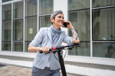 Yetişkin bir yaşlı beyaz kadın dışarıda duruyor. Cep telefonu kullanıyor. Gündüz güvenli modern alternatif ulaşım ve iletişim alanı kopyalama yönteminde elektrikli scooterla konuşuyor.