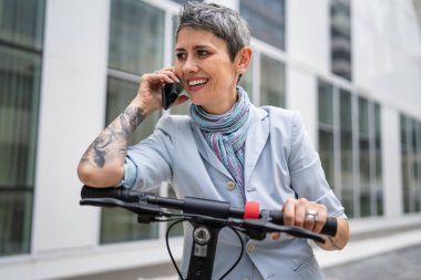 Yetişkin bir yaşlı beyaz kadın dışarıda duruyor. Cep telefonu kullanıyor. Gündüz güvenli modern alternatif ulaşım ve iletişim alanı kopyalama yönteminde elektrikli scooterla konuşuyor.