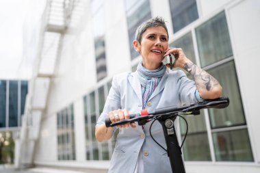 Yetişkin bir yaşlı beyaz kadın dışarıda duruyor. Cep telefonu kullanıyor. Gündüz güvenli modern alternatif ulaşım ve iletişim alanı kopyalama yönteminde elektrikli scooterla konuşuyor.