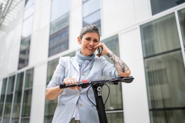 Yetişkin bir yaşlı beyaz kadın dışarıda duruyor. Cep telefonu kullanıyor. Gündüz güvenli modern alternatif ulaşım ve iletişim alanı kopyalama yönteminde elektrikli scooterla konuşuyor.