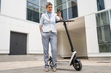 Yetişkin bir son sınıf beyaz kadın dışarıda duruyor selfie fotoğrafları için cep telefonu kullanıyor ya da elektrikli scooter 'ın güveni olan modern alternatif ulaşım aracı fotokopi uzay mesajları için video çağrısı yapıyor.
