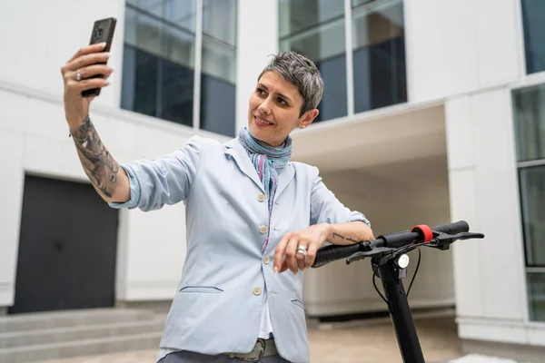 1人の女性成熟したシニア白人女性立って屋外使用の携帯電話のための自撮り写真やビデオ通話電動キックスクーターで一日に自信を持って現代的な代替モードの輸送コピースペース — ストック写真