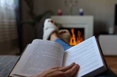 Bir kadın kış günü yatağında uzanıp şöminenin yanında kitap okuyor.