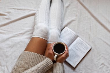 Evde bir kitapla, rahat bir yatakta yün çoraplar içinde, bacakları üzerinde kahve fincanı tutan bir kadın. Yaklaş, seçici odaklanma