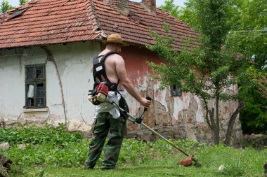 Çiftçi, bir yaz günü bahçede uzun çimleri, elleri petrol makası ile biçer. Bir adam güneşten korunmak için kafasına hasır şapka takar.