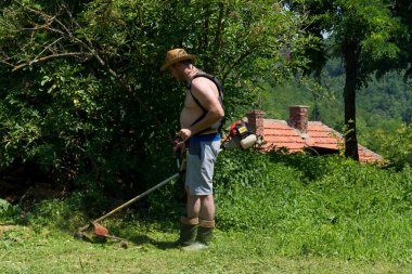 Hasır şapkalı çiftçi bir evin arkasındaki güneşli bir yaz gününde arka bahçede çim biçme makinesiyle uzun çimleri biçti. Bahçe konsepti
