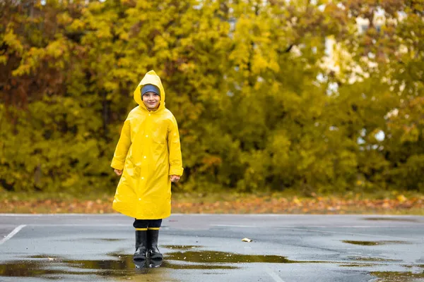 Dreng i gul vandtæt kappe og sorte gummistøvler går i en vandpyt i parken i regnvejr om efteråret. Udendørs aktivitet - Stock-foto