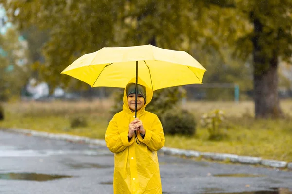 Sarı su geçirmez pelerinli ve şemsiyeli siyah lastik çizmeli bir çocuk sonbaharda yağmurda parkta yürüyor. Açık hava aktivitesi — Stok fotoğraf