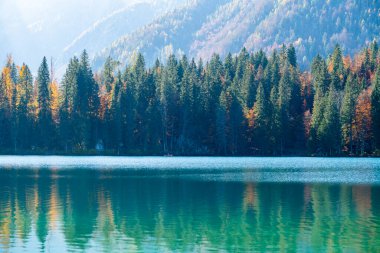 Dağ gölü, sonbahar ağaçları ve güneş ışığındaki güçlü dağlar.