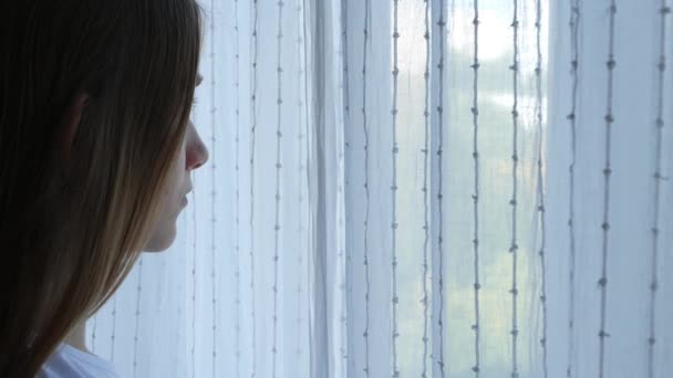 コロナウイルスに隔離された病気の悲しい女の子窓のカーテンを開けて、ガラス、退屈した子供を屋外で見る不幸な子供 — ストック動画