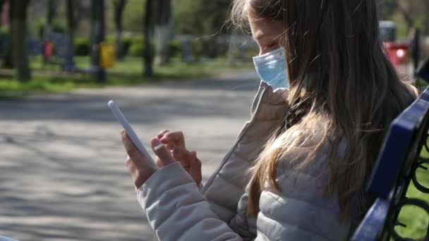在智能手机上玩保护面具的生病儿童，在公园的长椅上使用智能手机的悲伤女孩，在大流行病中的儿童 — 图库视频影像