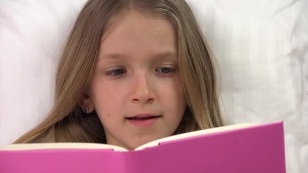 Niño sonriendo mientras lee un libro, niño relajándose en la cama en casa, riéndose cara de niña descansando en el dormitorio, educación infantil — Vídeo de stock