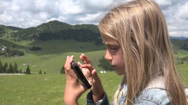 Enfant jouant Smartphone extérieur, L'enfant utilise une tablette dans les montagnes, Fille joue à des jeux en ligne sur dispositif de téléphone intelligent sur prairie dans la nature — Video