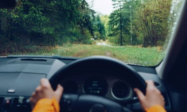 Sürücü parlak turuncu ceketli, modern bir off-road kullanıyor. Sağ el, dağ yeşil orman yolunda RHD araba sürüyor. POV arabanın ön camına bakıyor. Güvenli otomatik sürüş konsepti.