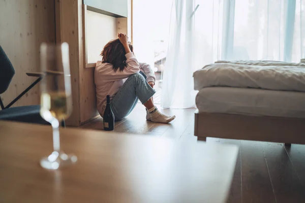 一个可悲的女人坐在卧室的地板上 喝着一瓶酒 没有聚焦的酒杯放在前排桌子上 心理健康和酗酒问题概念形象 — 图库照片