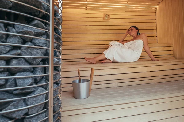 Relaxing Woman wrapped white towel lying and sweating on the wooden bench in Hot Finnish sauna with hot stones and enjoying pleasant body care temperature treatment. Healthy people lifestyle concept.