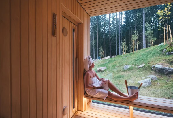 芬兰火热桑拿的木制长椅上 裹着白毛巾的松驰女人 有着广阔的窗户 绿树成荫 享受着令人愉快的降体温护理 — 图库照片