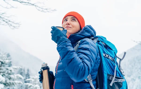 中年の女性は 冬の山岳ルートをトレッキングしながら 暖かいダウンジャケットを着て魔法瓶フラスコから熱い飲み物を笑って飲んでいます 自然コンセプトのイメージで活躍する人たち — ストック写真