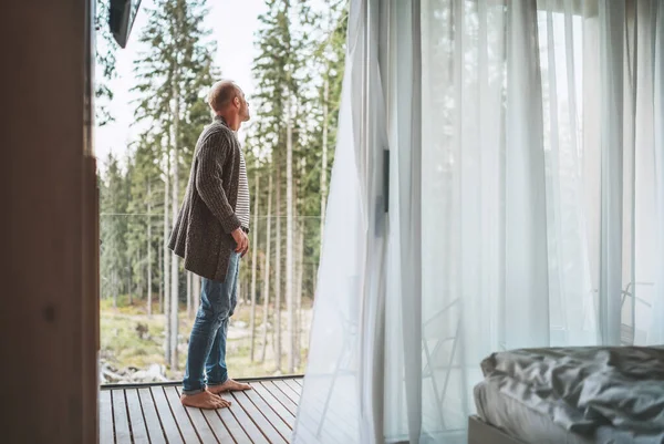 一名男子穿着敞开的针织羊毛衫 站在林屋阳台上 与自然森林松树一起享受新鲜空气 斯堪的纳维亚室内设计屋外面在野外生活的概念 — 图库照片