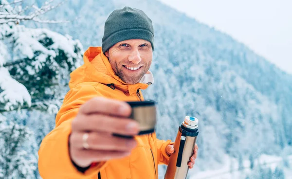 冬の山道をトレッキングしながら 明るいオレンジのソフトシェルジャケットを着た魔法瓶のフラスコから熱いドリンクカップを提供する中年の男 自然コンセプトのイメージで活躍する人たち — ストック写真