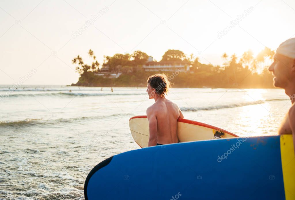 Young teen boy with a surfboard with his father go to the sea for surfing. They are smiling and enjoying a beautiful sunset light. Family active vacation concept.