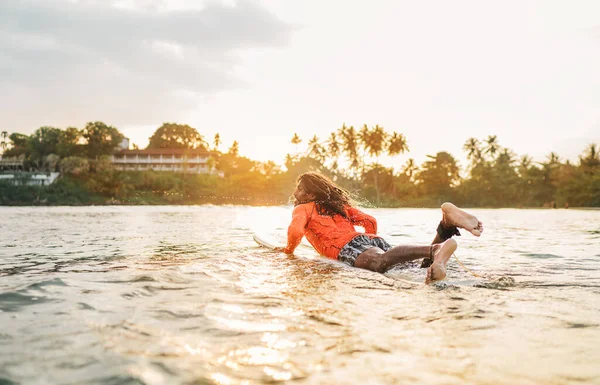 一头乌黑的长发男子坐着长长的冲浪板划向印度洋的冲浪点 棕榈树在背景中闪烁着落日的光芒 极端水上运动和到异国他乡旅游的概念 — 图库照片