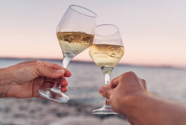 Birbirlerine aşık çiftler, güzel pembe deniz kenarında gün batımının tadını çıkarırken beyaz şarap kadehleriyle şıngırdarlar. Romantik tatil ya da flört konsepti yakın plan görüntü.