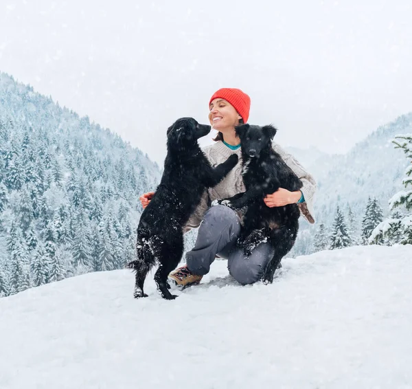 一个穿着保暖衣服的年轻女子在一座风景如画的雪山户外遛狗 雌性笑着和宠物玩耍 还有一只狗舔主人的下巴 人类和宠物冬季的概念形象 — 图库照片