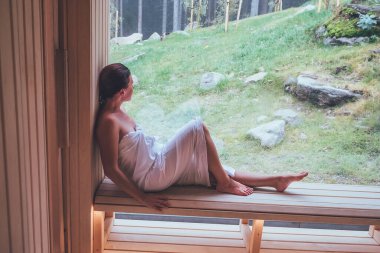 Ateşli Finlandiya saunasındaki ahşap bankta oturan, yeşil orman manzaralı geniş pencereli ve hoş, sağlıklı vücut sıcaklığı tedavisinin keyfini çıkaran, rahatlatıcı beyaz kadın..