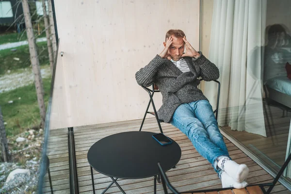 一个疲惫的中年男人穿着开襟羊毛衫 牛仔裤和温暖的袜子坐在林屋阳台上的画像 他按摩太阳穴以增加血液循环和缓解头痛 — 图库照片