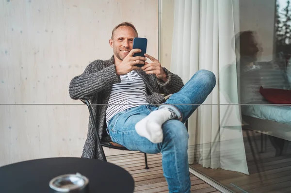 笑着的中年男人穿着开著的羊毛衫 牛仔裤和温暖的袜子坐在房子的阳台上 当他使用现代智能手机时 他笑了 每天的生活方式照片与现代装置的概念形象 — 图库照片