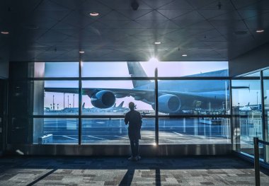 Yalnız gezgin çocuk havaalanı terminalinde geniş bir pencerenin yanında duruyor ve sabah güneşiyle uçuşa hazırlanan uçağa binmek için bekliyor..