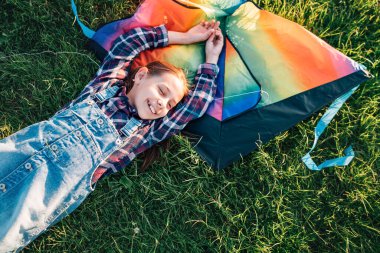 9YO gülümseyen kız renkli gökkuşağı uçurtma oyuncağıyla çimlerin üzerinde yatıyor. Mutlu çocukluk anları ya da açık hava harcama konsepti resmi.