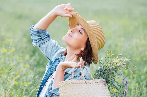 一个年轻女人的画像闭上了眼睛 欣赏着阳光 她穿着牛仔裤夹克 草帽和浅色夏装 她在草地上摘了一朵野花 把花束放在篮子里 — 图库照片