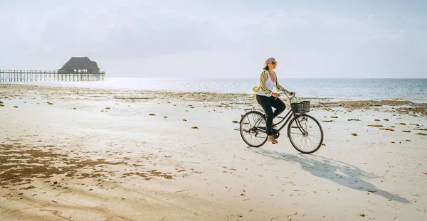若い女性は タンザニアのザンジバル島のKiwengwaビーチの孤独な干潮の海の白い砂の海岸にフロントバスケットで古いヴィンテージの自転車に乗って軽い夏の服を着て — ストック写真