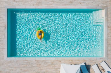 Siyah bikinili seksi bir kadın, hasır şapkalı ve güneş gözlüklü mavi yüzme havuzunda yüzen dev sarı ananas tüplü. Lüks tatil beldelerinde yaz tatili konsepti..