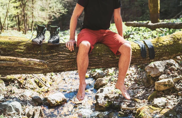 男は靴下の洗濯乾燥やトレッキングブーツを待っている間 渓流の上の倒木のログに座っています アクティブな人々の旅行 ハイキングやトレッキングのコンセプトイメージ — ストック写真